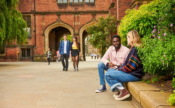 Two people sitting on a stone bench talking on campus. Behind them, others walk along a path leading to the Arches, surrounded by trees and greenery.